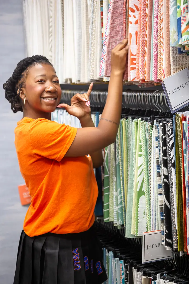 Graduate laughing while reaching for a hanging fabric sample in a design showroom.